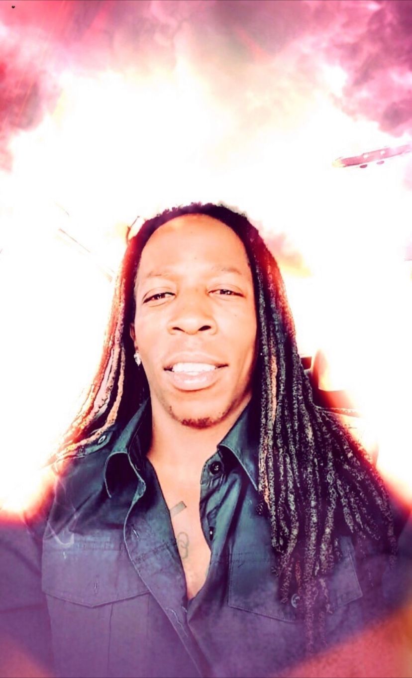 Man with long dreadlocks, wearing a dark shirt, smiling, against a cloudy, bright background.