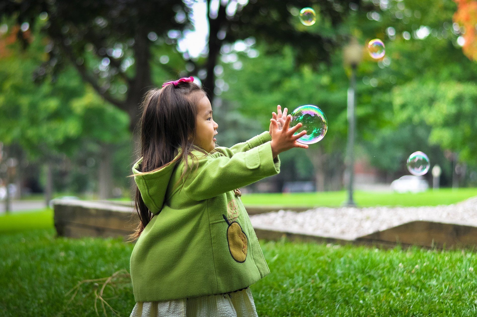 Girl in green coat reaching for bubbles in a park.