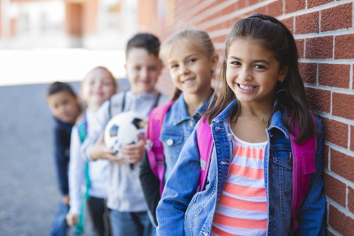 Children with backpacks smile, leaning against a brick wall. One holds a soccer ball.