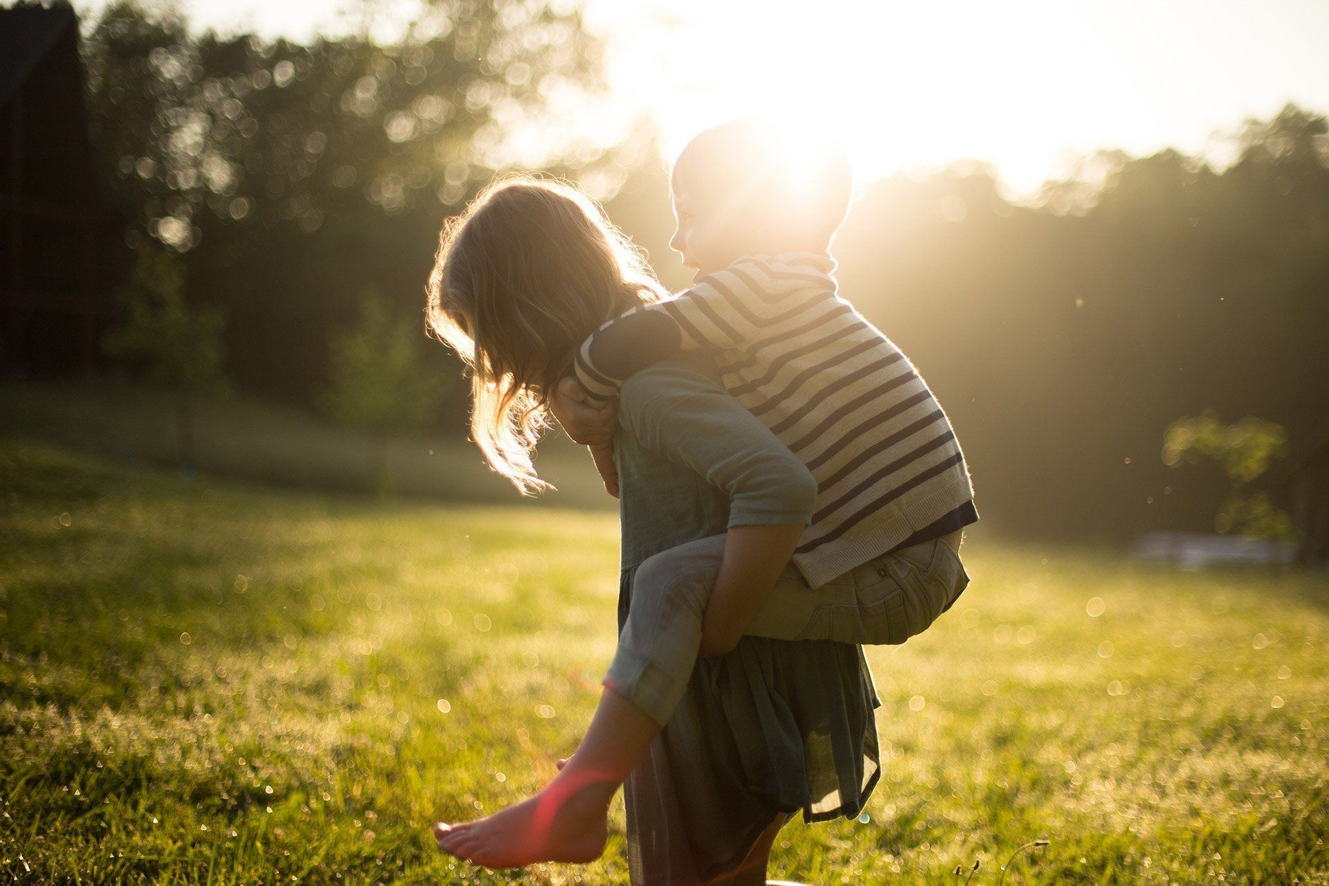 Girl carrying a child piggyback-style in a sunny grassy field.