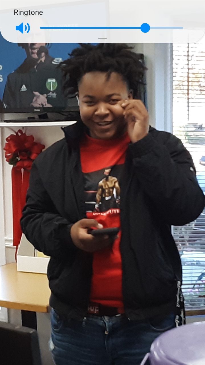 Person smiling, holding phone, wearing black jacket over red shirt with graphic, in front of a shelf with decor.