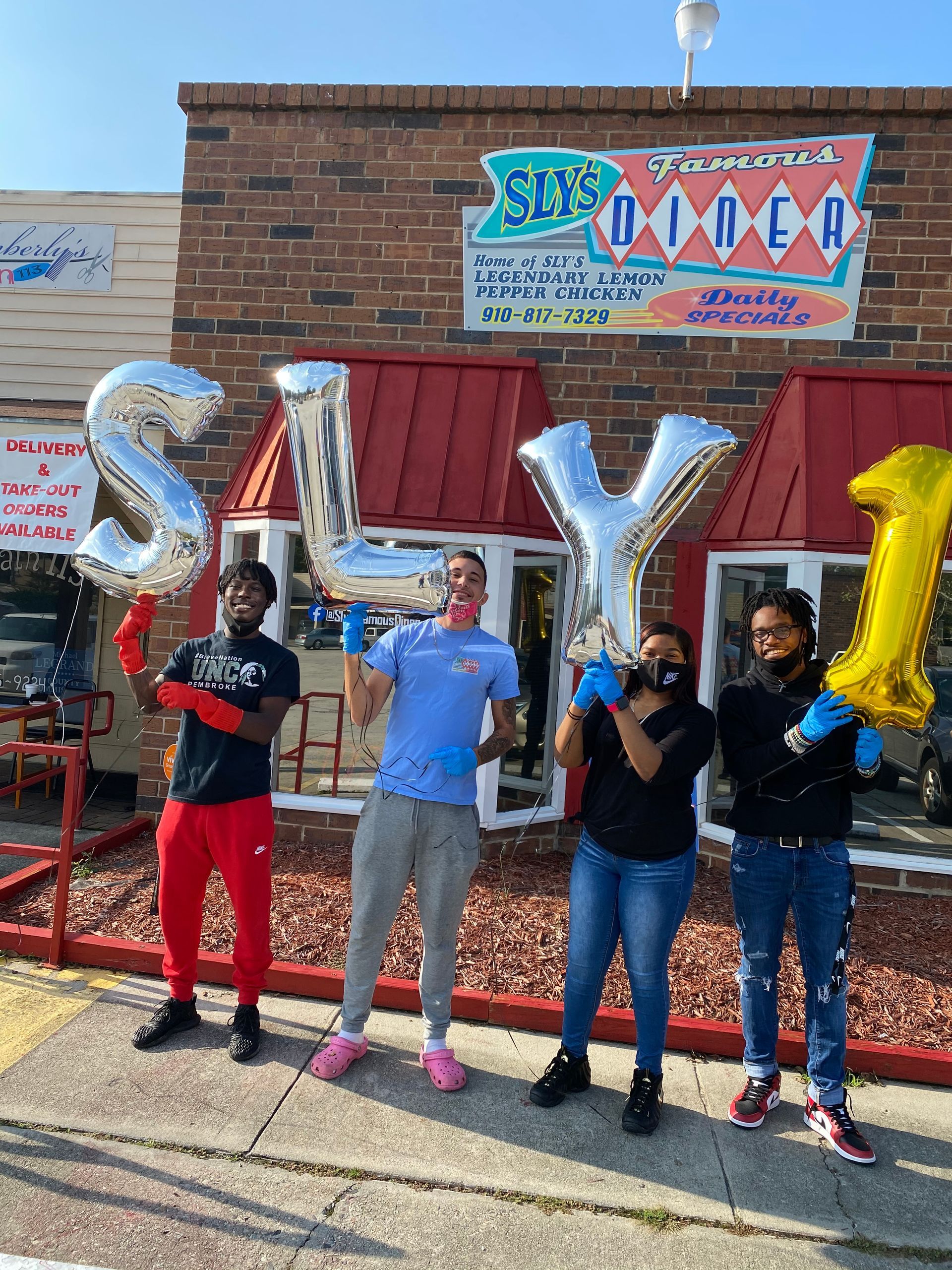 Four people outside Sly's Diner holding balloon letters spelling 